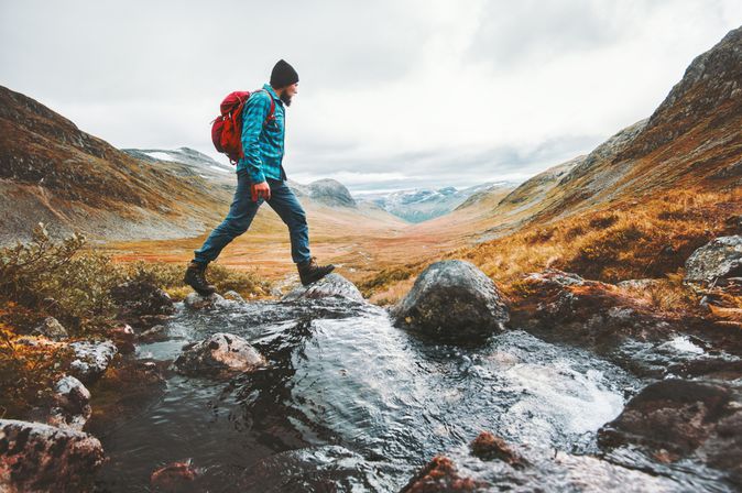 Ein Mann in Wanderkleidung läuft in einer Berglandschaft über Steine in einem Bach
