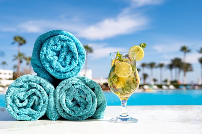 Drei blaue aufgerollte Handtücher liegen am Poolrand, daneben ein gelbes Getränk mit Zitrone