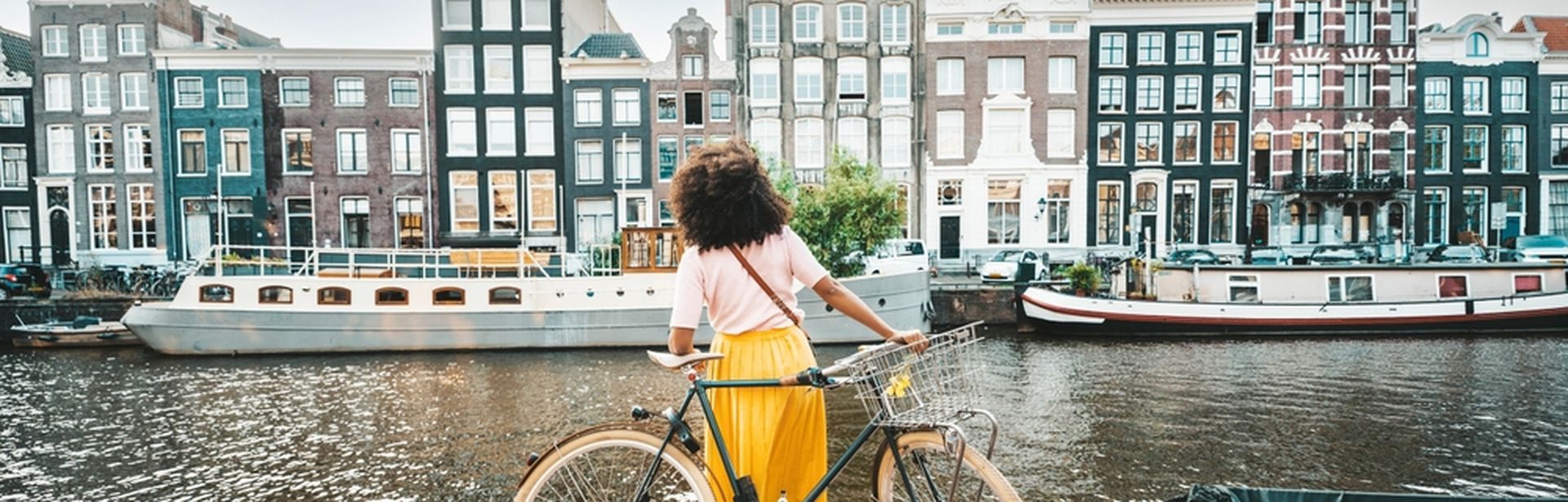 Eine Frau in einem gelben Rock steht an ihrem Fahrrad und schaut auf die Amstel und die gegenüberliegenden Häuser und Boote in Amsterdam
