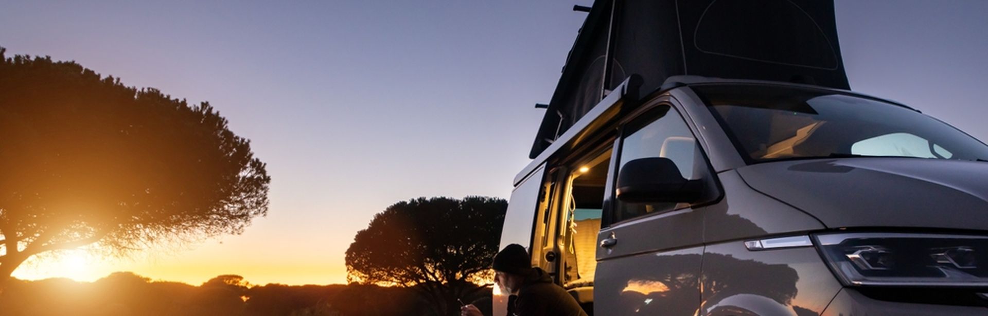 Ein Mann sitzt im Eingang eines Transporters und schaut auf sein Smartphone, im Hintergrund große Bäume und ein Sonnenuntergang