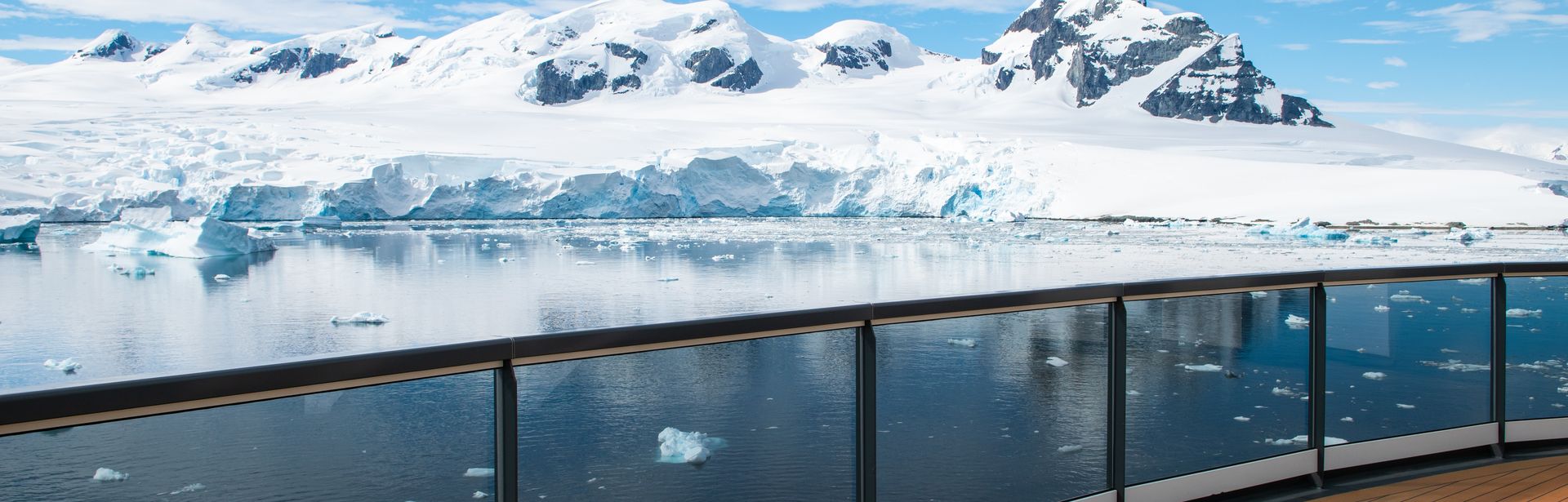 Ein Schiffsdeck mit Glasgeländer bietet einen Blick auf eine antarktische Landschaft mit schneebedeckten Bergen und schwimmenden Eisschollen