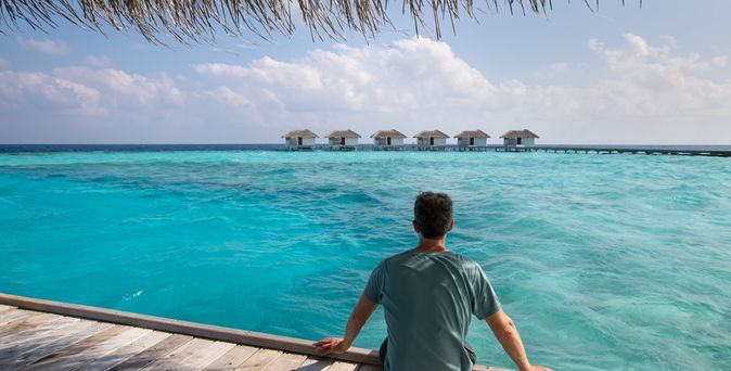 Ein Mann sitzt auf einem Steg am Meer und schaut auf mehrere Wasserbungalows