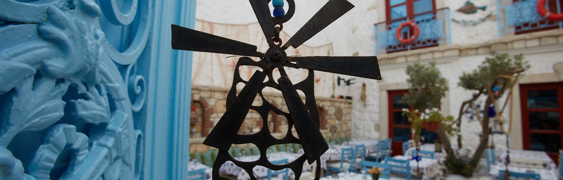 Ein Windspiel in Form einer Windmühle hängt in einem Hinterhof mit bunt gedeckten Tischen und blauen und weißen Stühlen