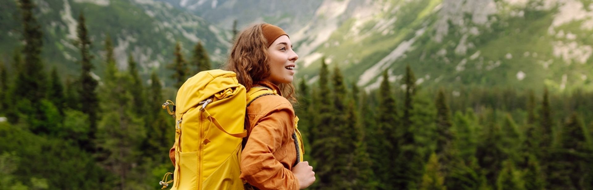 Eine Frau mit gelbem Wanderrucksack steht auf einem Berg und schaut begeistert in die Ferne