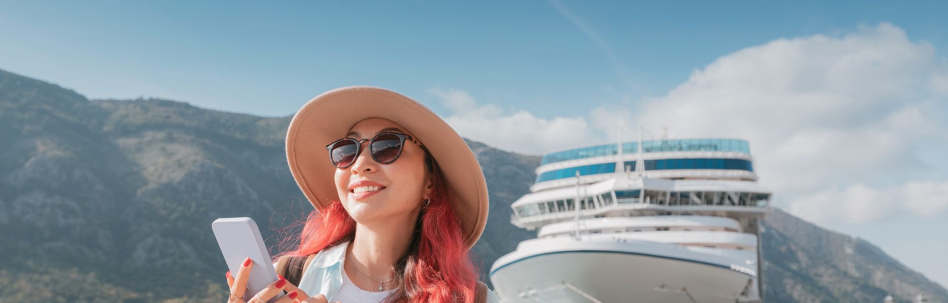 Frau mit Rosa Haaren und Handy in der Hand steht am Hafen vor einem Kreuzfahrschiff