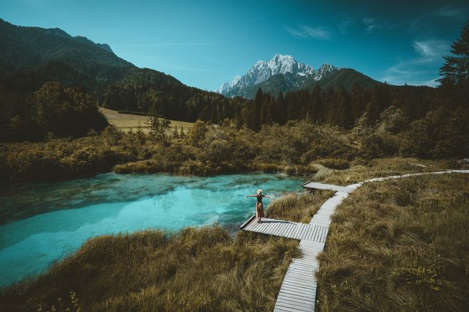 Eine Große Wiese mit einem Holzweg, auf einem Steg steht eine Frau und breitet die Arme aus, sie schaut auf den See vor ihr