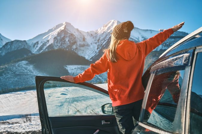 Frau steht an der offenen Tür eines Autos, blickt auf eine schneebedeckte Berglandschaft und streckt die Arme aus