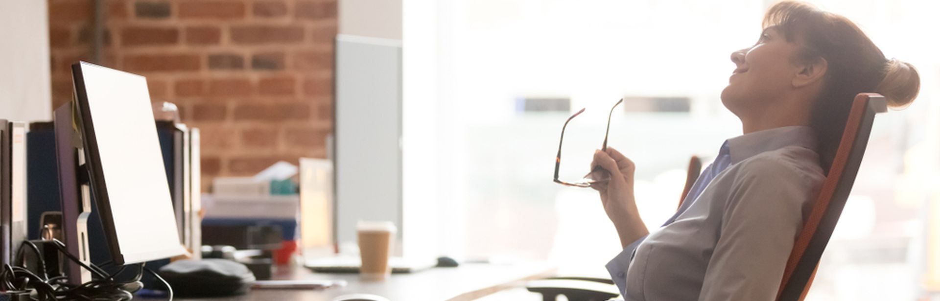Eine Frau sitzt zurückgelehnt an einem Schreibtisch und hält eine Brille in der Hand