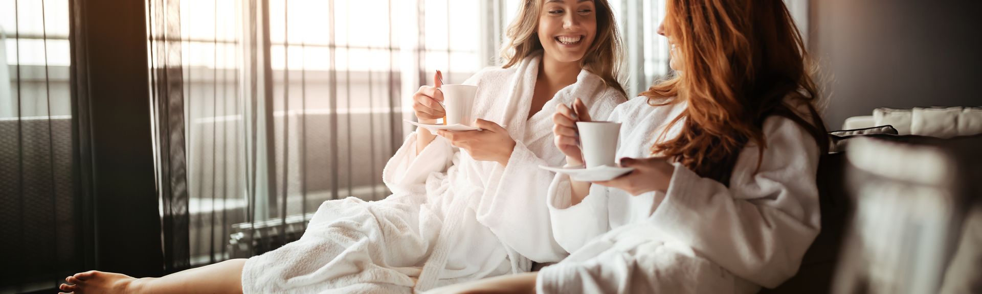 Zwei lachende Frauen sitzen in Bademänteln auf einem Sofa und Trinken Tee