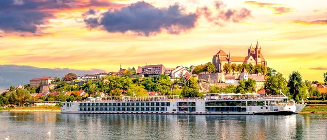 Langes Flusskreuzfahrschiff fährt entlang eines kleinen Dorfes, bei gelb leuchtendem Himmel