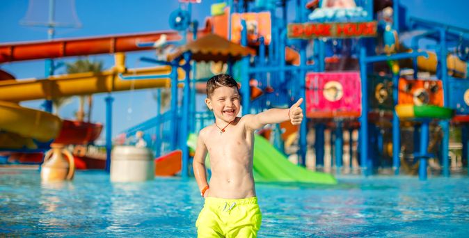 Ein Junge in einer gelben Badehosen steht vor einem großen Aquapark und zeigt einen Daumen nach oben