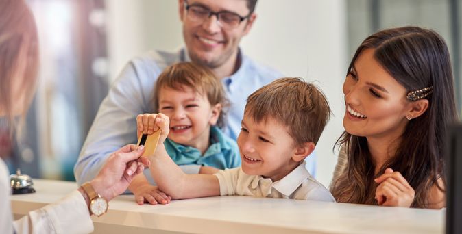 Eine Familie mit zwei Kindern beim Check-In, die Rezeptionistin reicht dem kleinen Kind die Zimmerkarte