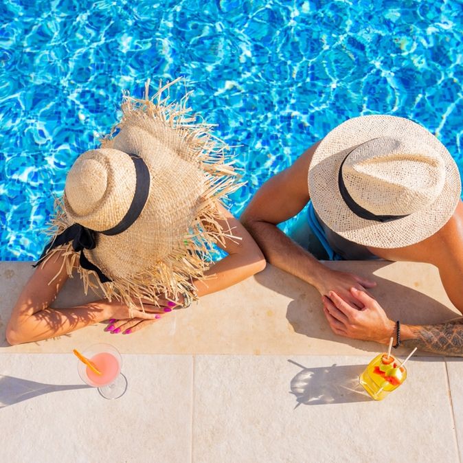 Zwei Personen von oben fotografiert, Sie tragen Strohhüte und man sieht Ihre Gesichter nicht. Sie sind in einem Pool und lehnen am Rand, vor Ihnen stehen Getränke auf dem Boden