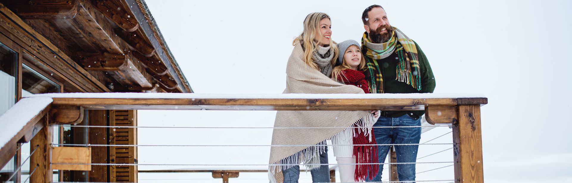Eine Familie steht in Decken gehüllt auf dem Balkon Ihres Hotels im Schnee