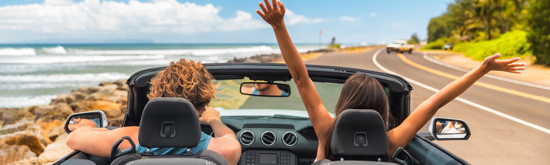 Zwei Personen im Auto mit offenem Verdeck, im Hintergrund kann man das Meer sehen