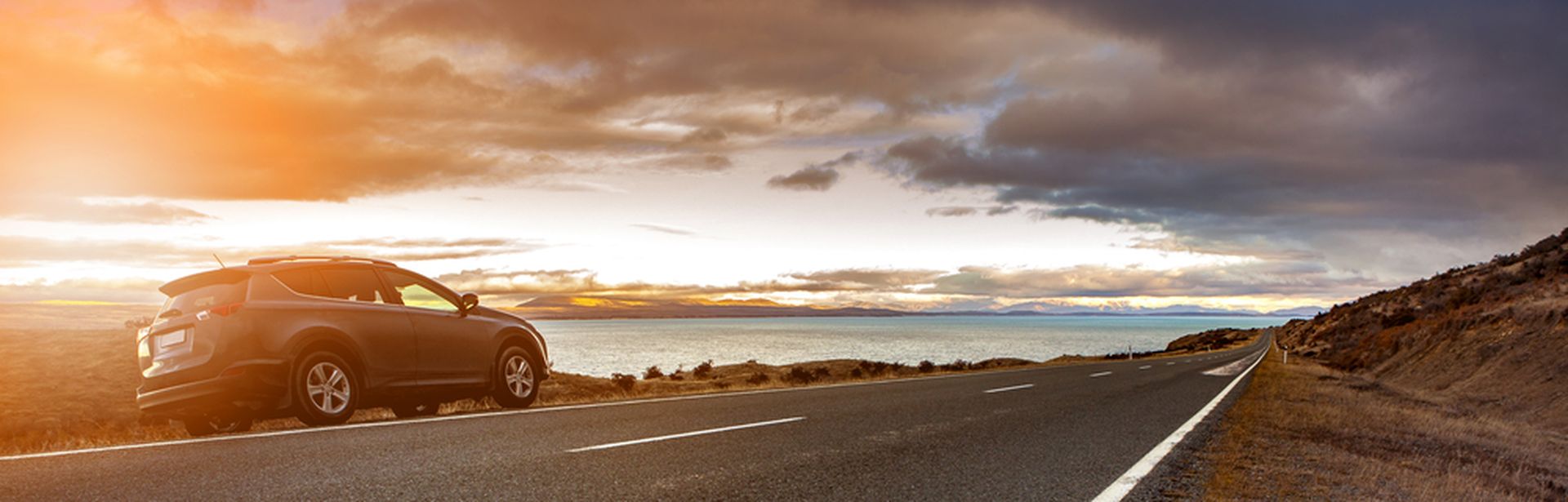 Ein Auto fährt bei Sonnenaufgang an der Kamera vorbei auf einer Straße am Wasser.