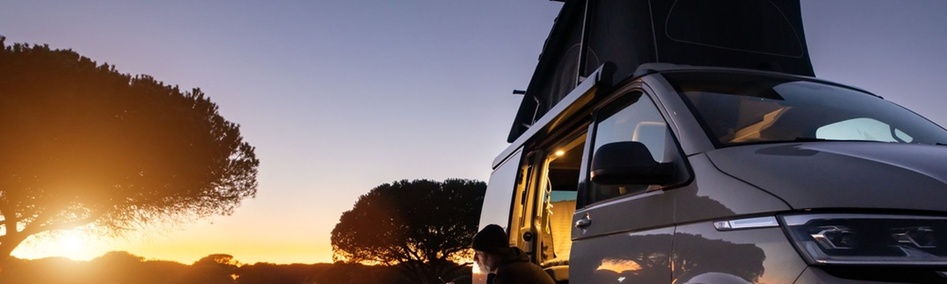 Ein Mann sitzt im Eingang eines Transporters und schaut auf sein Smartphone, im Hintergrund große Bäume und ein Sonnenuntergang