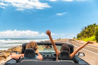 Zwei Personen fahren in einem Auto eine Straße am Meer entlang, eine von ihnen reißt die Arme nach oben
