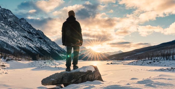 Mann steht leicht erhöht auf einem großen Stein in den Bergen bei Schnee und schaut in die untergehende Sonne