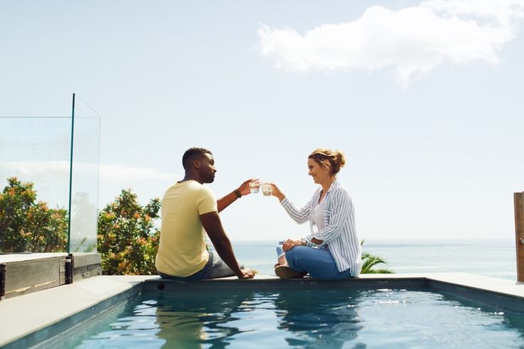 Eine Frau und ein Mann sitzen am Pool und stoßen mit Ihren Getränken an