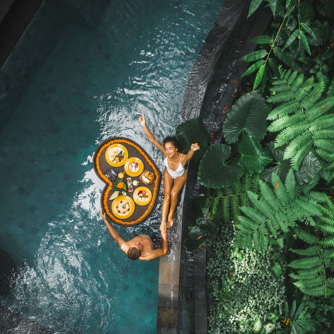 Ein Paar von oben, eine Frau sitzt am Rand eines Pools, ein Mann steht im Pool, zwischen ihnen schwimmt ein Herzförmiges Tablett mit Essen