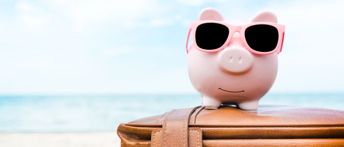 Ein rosanes Sparschwein mit Sonnenbrille sitzt auf einem Koffer am Strand