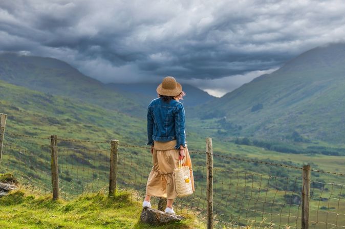 Frau steht auf einem Stein und blickt auf auf eine grüne Berglandschaft