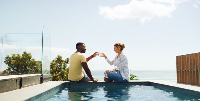 Eine Frau und ein Mann sitzen am Pool und stoßen mit Ihren Getränken an