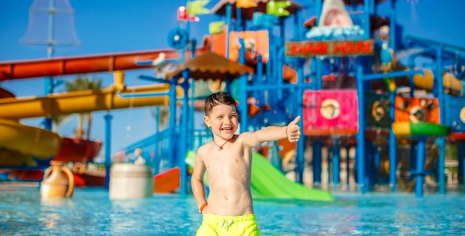 Ein Junge in einer gelben Badehosen steht vor einem großen Aquapark und zeigt einen Daumen nach oben