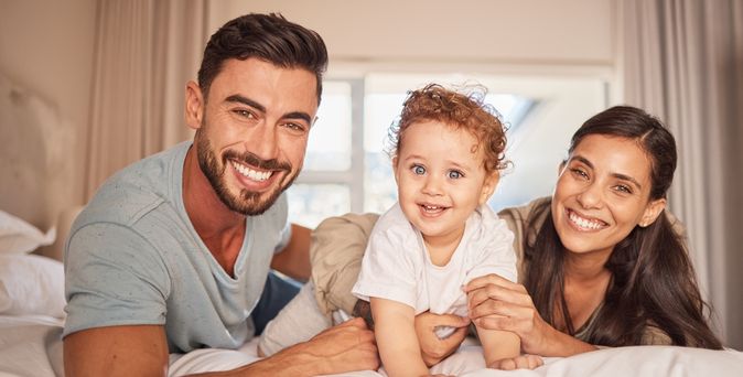 Eltern mit ihrem Kind liegen auf dem Bett und lachen in die Kamera