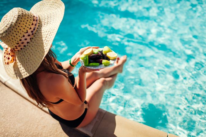 Eine Frau von oben fotografiert, sie trägt einen Strohhut und sitzt im Bikini am Pool, in der Hand hat sie einen Teller mit geschnittenem Obst