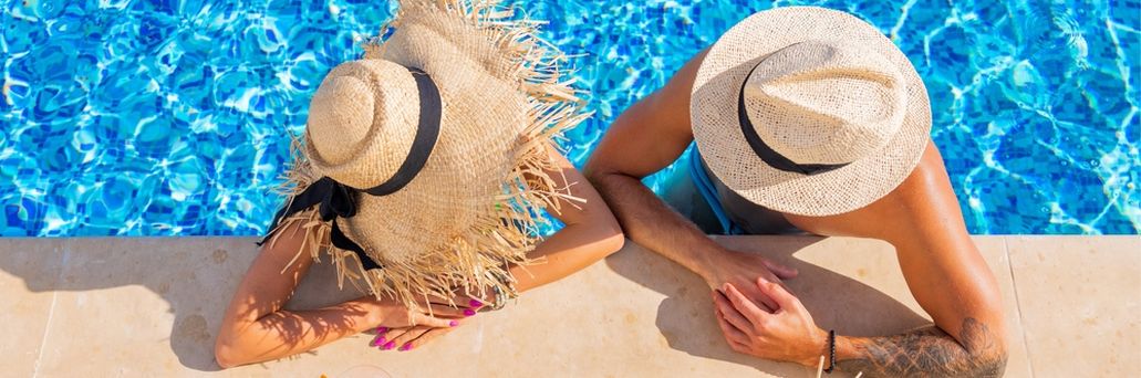 Zwei Personen von oben fotografiert, Sie tragen Strohhüte und man sieht Ihre Gesichter nicht. Sie sind in einem Pool und lehnen am Rand, vor Ihnen stehen Getränke auf dem Boden