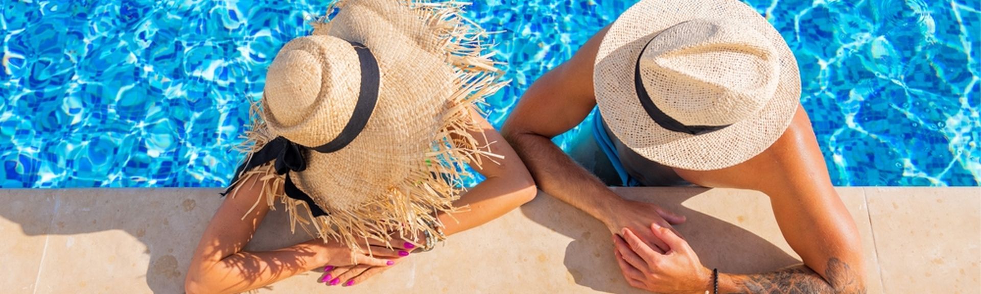 Zwei Personen von oben fotografiert, Sie tragen Strohhüte und man sieht Ihre Gesichter nicht. Sie sind in einem Pool und lehnen am Rand, vor Ihnen stehen Getränke auf dem Boden