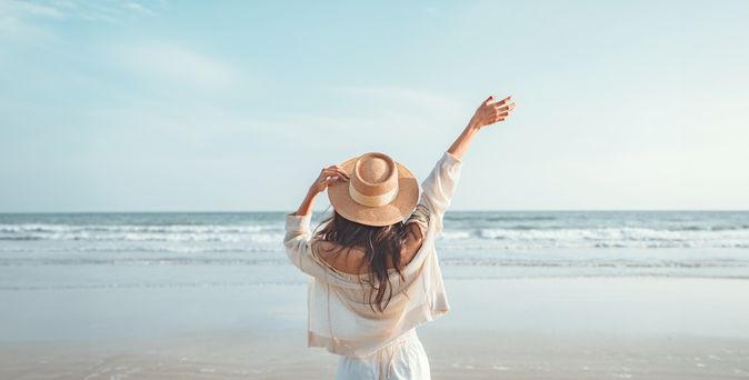 Eine Frau mit Hut von hinten, Sie schaut vom Strand aus auf das Meer und hebt den Arm 