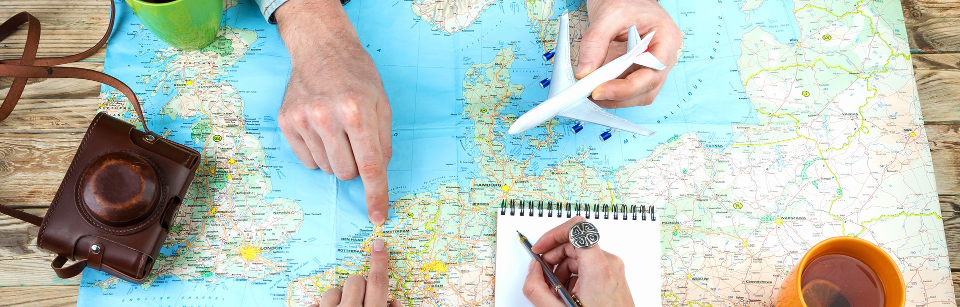 Weltkarte auf Holztisch ausgebreitet, Zwei Personen zeigen mit Fingern auf verschiedene Orte auf der Karte, Eine Person hält ein Modellflugzeug, Notizblock mit Stift, Zwei Tassen stehen auf dem Tisch, Alte Kamera mit Ledertasche