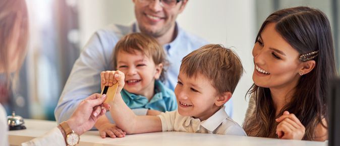 Eine Familie mit zwei Kindern beim Check-In, die Rezeptionistin reicht dem kleinen Kind die Zimmerkarte