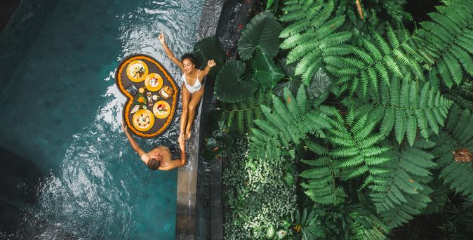 Ein Paar von oben, eine Frau sitzt am Rand eines Pools, ein Mann steht im Pool, zwischen ihnen schwimmt ein Herzförmiges Tablett mit Essen