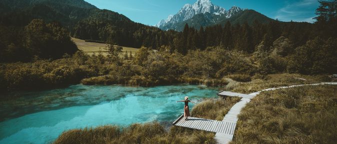 Eine Große Wiese mit einem Holzweg, auf einem Steg steht eine Frau und breitet die Arme aus, sie schaut auf den See vor ihr