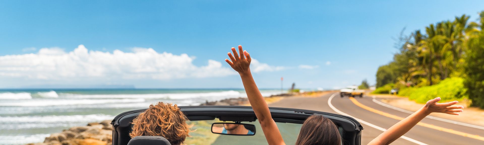 Zwei Personen im Auto mit offenem Verdeck, im Hintergrund kann man das Meer sehen