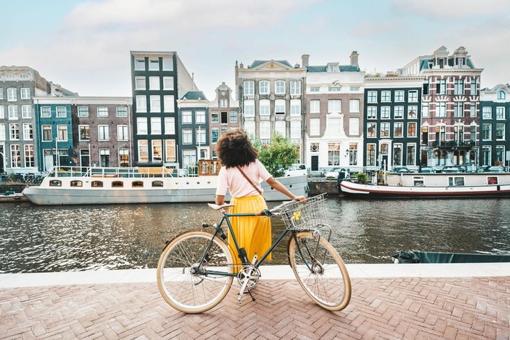 Eine Frau in einem gelben Rock steht an ihrem Fahrrad und schaut auf die Amstel und die gegenüberliegenden Häuser und Boote in Amsterdam