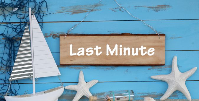 Eine blaue Holzwand mit einem Schild mit der Aufschrift "Last Minute", davor stehen zwei Seesterne, ein kleines Holzboot und eine kleine Flasche mit Muscheln und Sand 