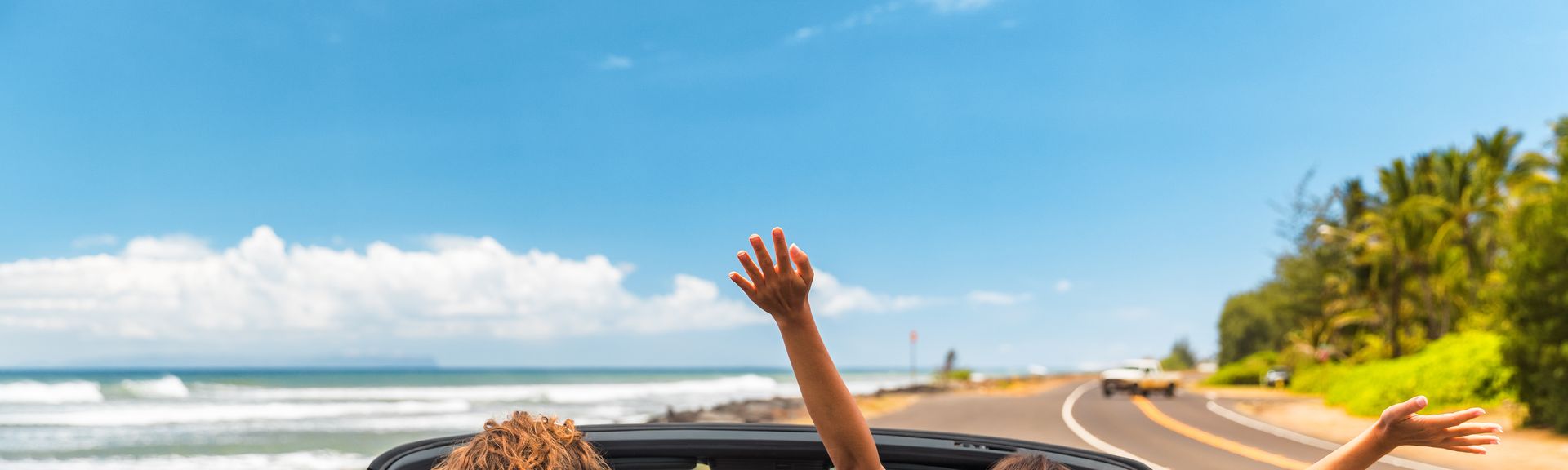 Zwei Personen im Auto mit offenem Verdeck, im Hintergrund kann man das Meer sehen