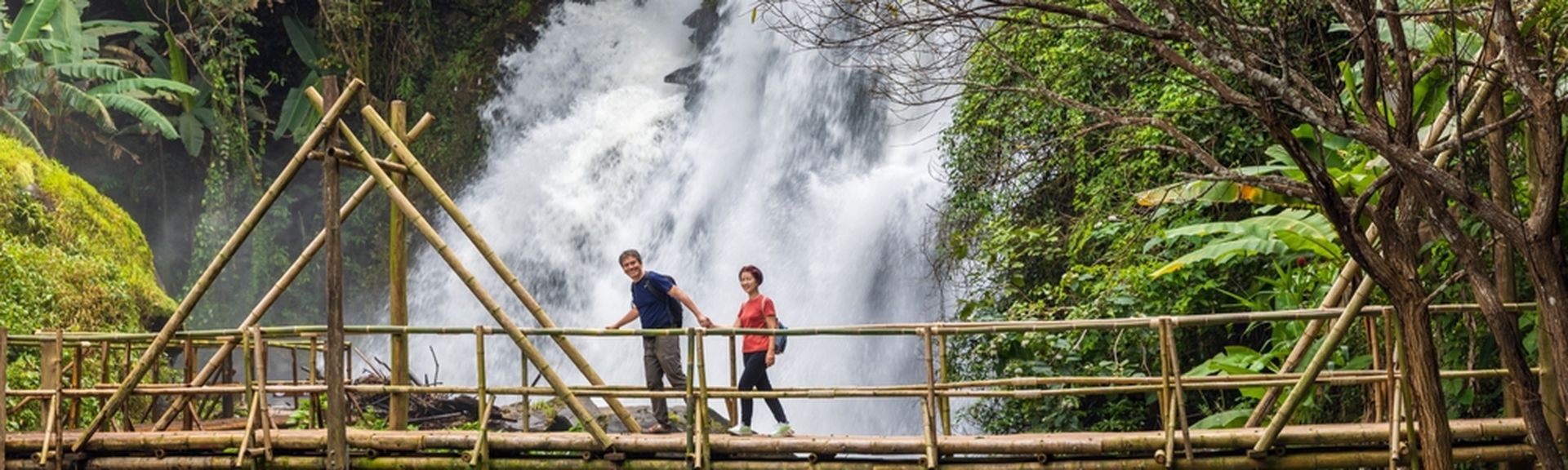 Eine Frau und ein Mann laufen Hand in Hand über eine Holzbrücke über einem Fluss, im Hintergrund ist ein großer Wasserfall