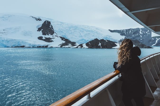 Eine Frau steht mit wehenden Haaren an der Reling eines Kreuzfahrtschiffes und schaut auf schneebedeckte Berge
