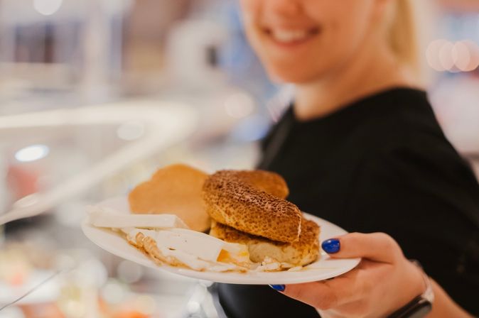 Eine lächelnde Frau hält einen Teller mit Brötchen, Käse und anderem Frühstück in die Kamera
