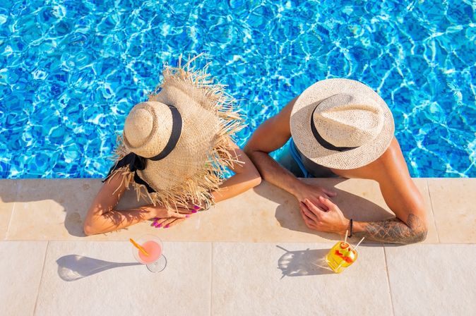 Zwei Personen von oben fotografiert, Sie tragen Strohhüte und man sieht Ihre Gesichter nicht. Sie sind in einem Pool und lehnen am Rand, vor Ihnen stehen Getränke auf dem Boden