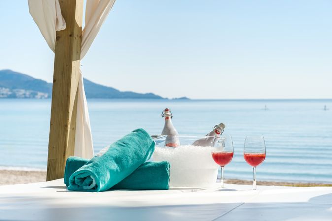 Zwei aufgerollte Handtücher liegen neben zwei Gläsern und ein Getränkekühler mit Eis und zwei Flaschen auf einer Decke am Strand