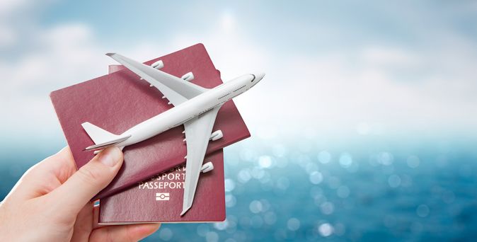 Eine Hand hält vor einem blauen Hintergrund zwei Reisepässe und ein Miniaturflugzeug hoch