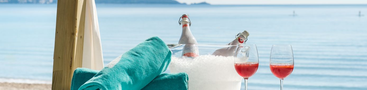 Zwei aufgerollte Handtücher liegen neben zwei Gläsern und ein Getränkekühler mit Eis und zwei Flaschen auf einer Decke am Strand