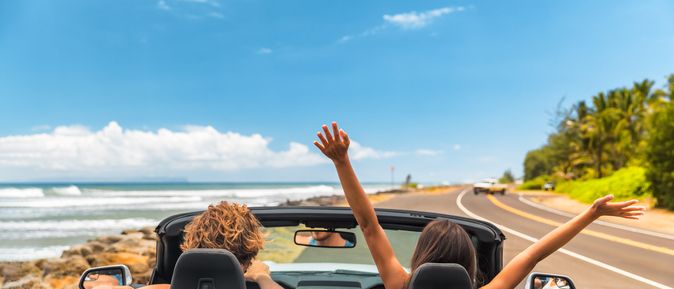 Zwei Personen im Auto mit offenem Verdeck, im Hintergrund kann man das Meer sehen